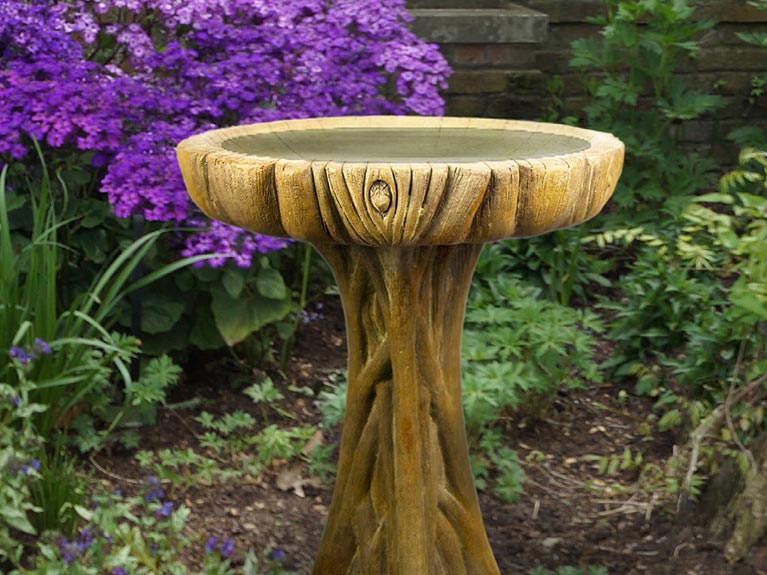 Stone bird bath in the shape of a tree trunk sitting outside near purple flowers