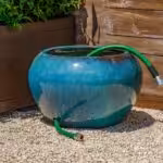 Large blue pottery planter sitting on gravel with holes for garden hose