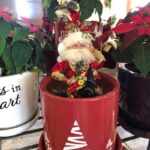 A small Mark Roberts Santa doll in a red flower pot with a white Christmas tree on it