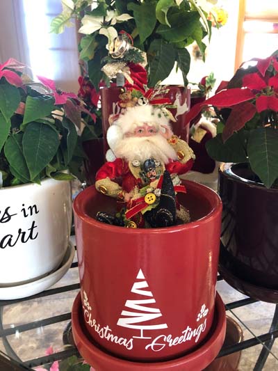 A small Mark Roberts Santa doll in a red flower pot with a white Christmas tree on it