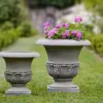 Two cast stone planter sitting on grass, one with purple flowers