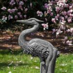 Garden statue of crane sitting on grass in front of purple tulip trees